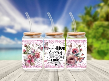 Drei 16 oz Glasbecher mit Bambusdeckel und Glastrinkhalm, verziert mit Blumenmotiven, Schmetterlingen und dem motivierenden Spruch 'Live every moment, laugh every day, love beyond words'. Die Becher stehen auf einem Holztisch vor einem tropischen Strandhi